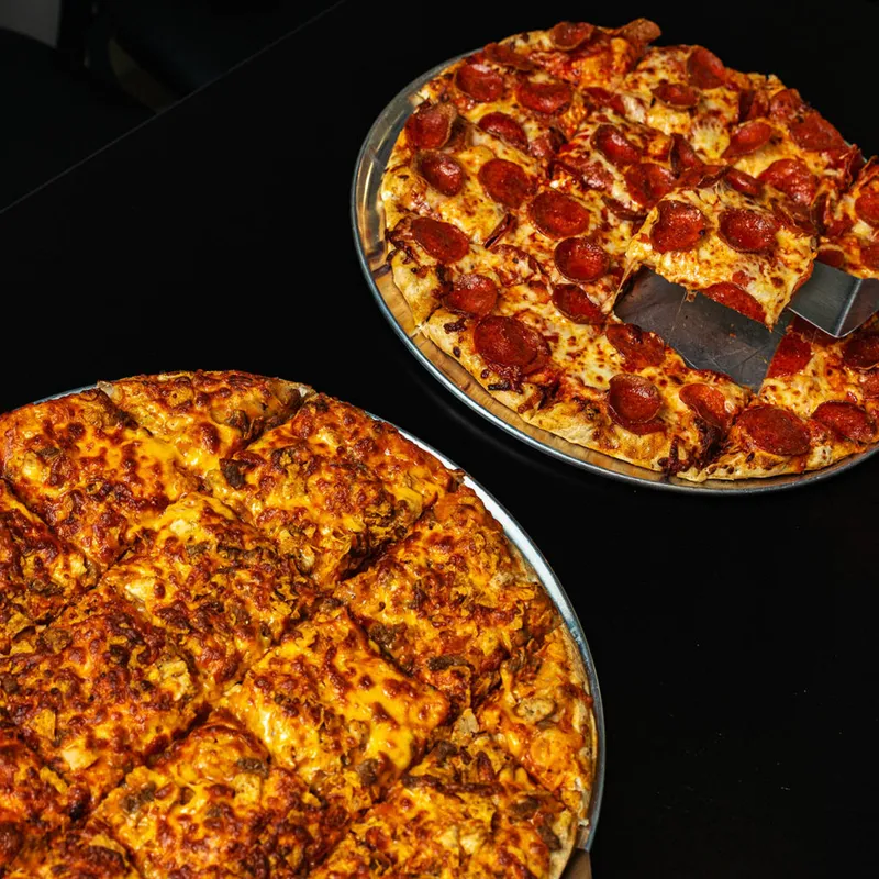 Two large pizzas on metal pans — one pepperoni, one cheesy vegetable — on a dark surface
