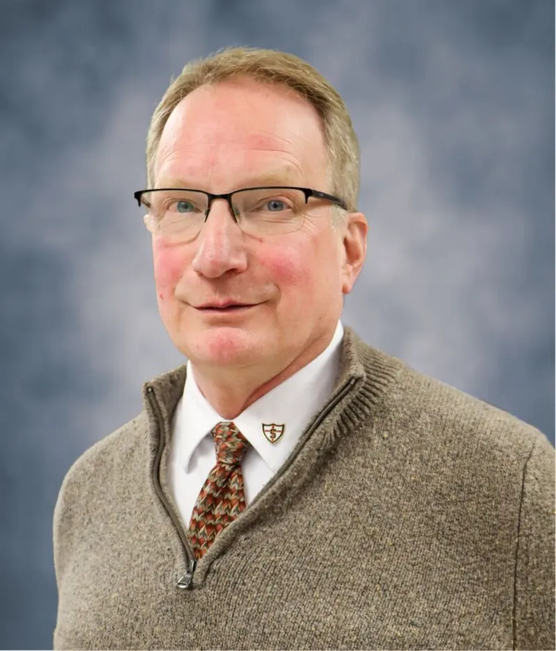 Middle-aged man with glasses wearing a tan zip sweater, dress shirt, and patterned tie against a blue backdrop
