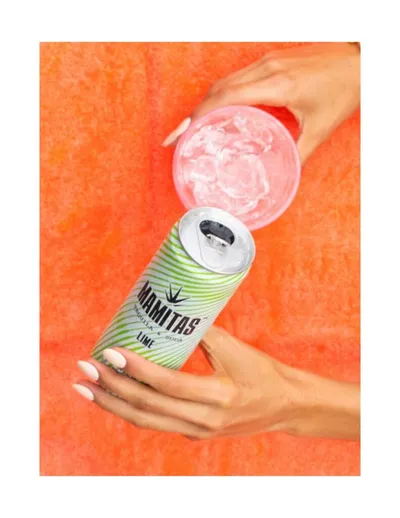 Hands holding a Mamitas lime canned cocktail and an iced pink drink against an orange background
