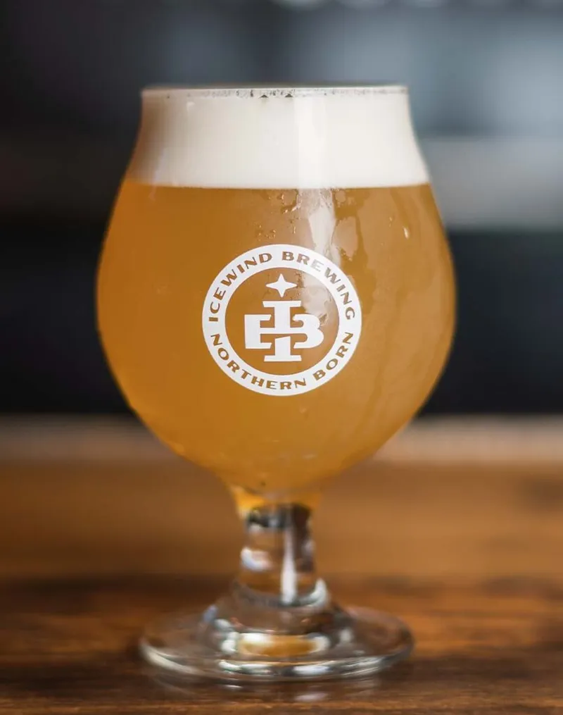 Tulip glass of hazy golden beer with white foam head, Icewind Brewing Northern Born logo etched on glass, on a wood bar