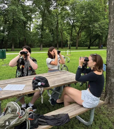 Three young people look through binoculars at a park picnic table surrounded by trees