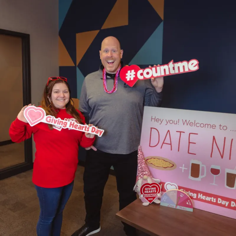Two people smiling and holding Giving Hearts Day signs beside a Date Night banner