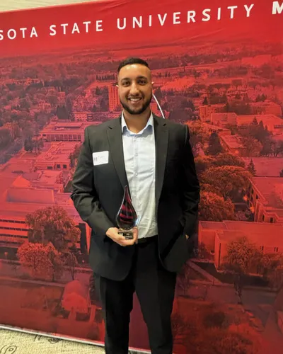 Man in dark suit holding a glass award in front of a red Minnesota State University banner