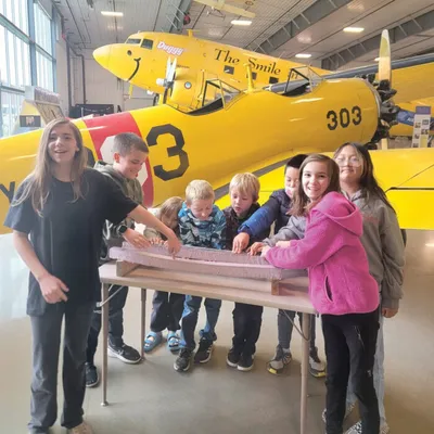 Children and adults gathered around a table exhibit inside a hangar with a yellow vintage aircraft behind them