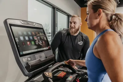 Bearded male trainer guides a woman on a treadmill with a touchscreen console in a gym