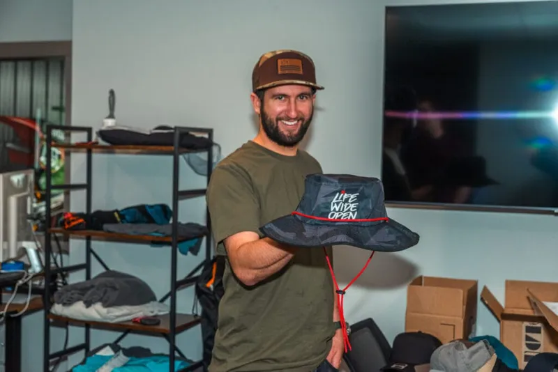 Smiling bearded man holding a dark bucket hat reading 'Life Wide Open' in a merchandise-filled room