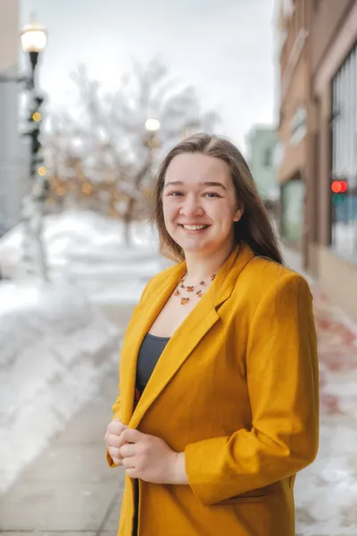 Young woman in a mustard blazer and necklace smiling on a snowy urban sidewalk