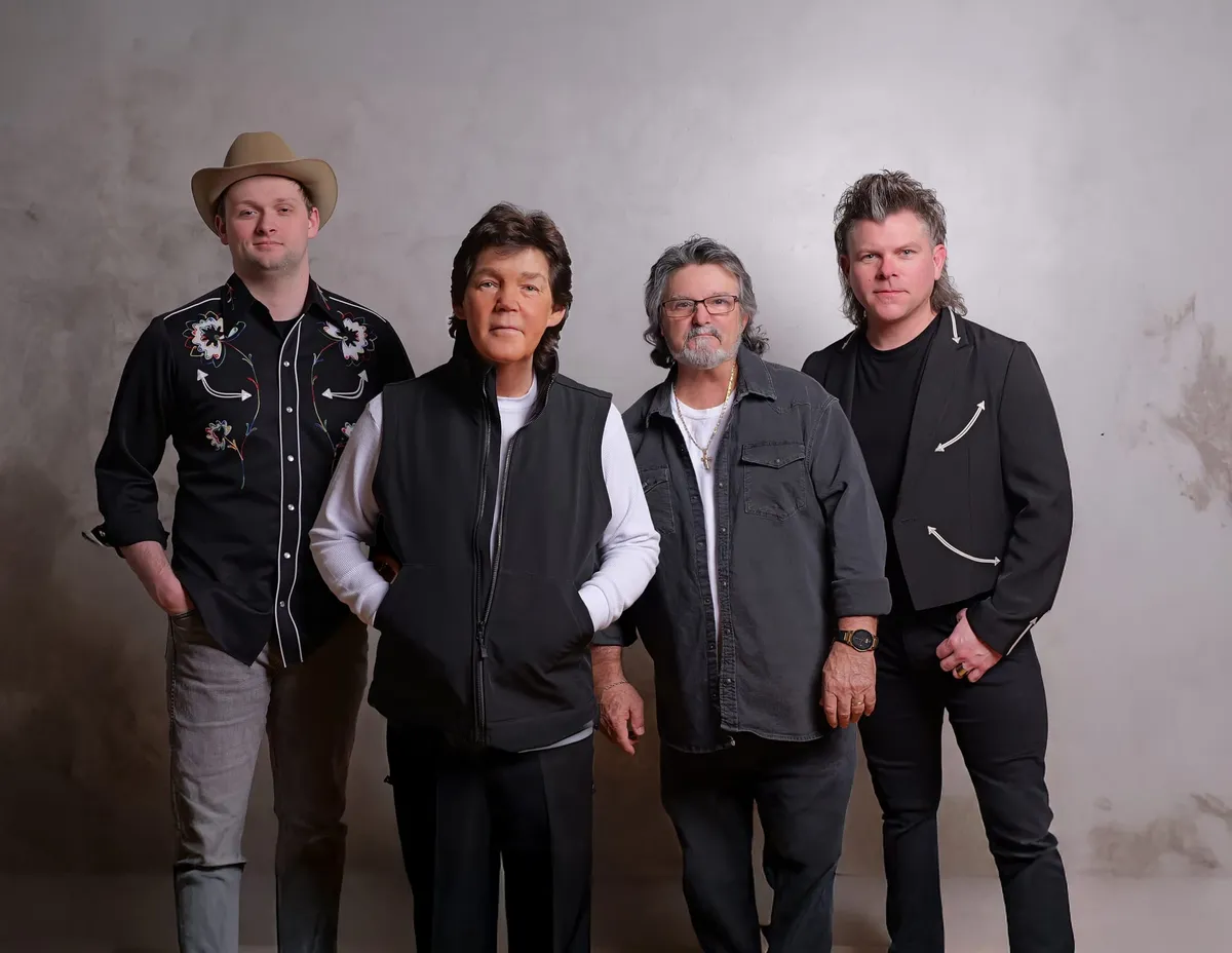Four men posing together against a gray studio backdrop, one wearing a tan cowboy hat and floral western shirt