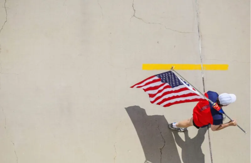 Aerial view of a runner in a red shirt carrying a waving American flag over concrete