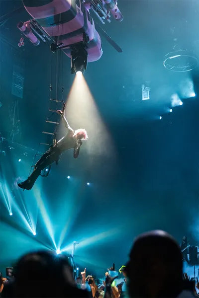 Pink-haired performer suspended mid-air by rigging above a packed concert crowd in blue stage lighting