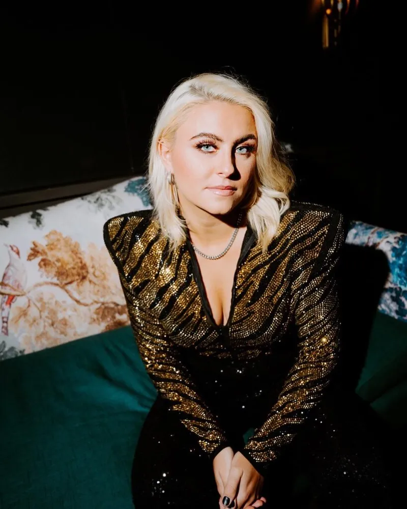 Blonde woman in a gold sequined blazer seated on a teal velvet sofa