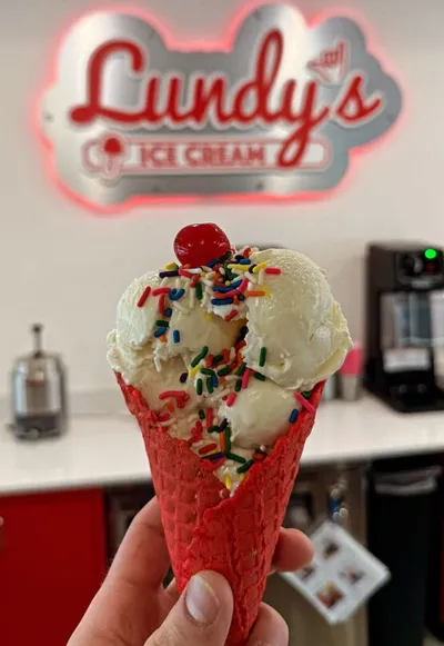 Two scoops of vanilla ice cream with rainbow sprinkles and a cherry on a red waffle cone, held in front of a Lundy's Ice