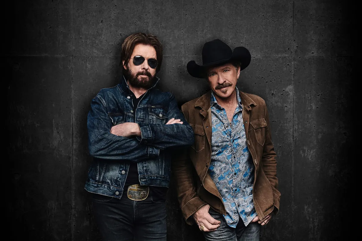 Two men in country western style lean against a dark wall, one in sunglasses and denim, one in a cowboy hat and suede jacket