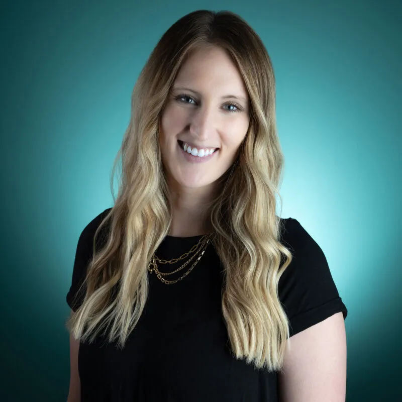 Smiling woman with long blonde hair in a black top and layered gold chain necklaces against a teal background