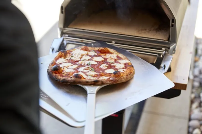 Charred Neapolitan-style pizza on a metal peel in front of an outdoor pizza oven