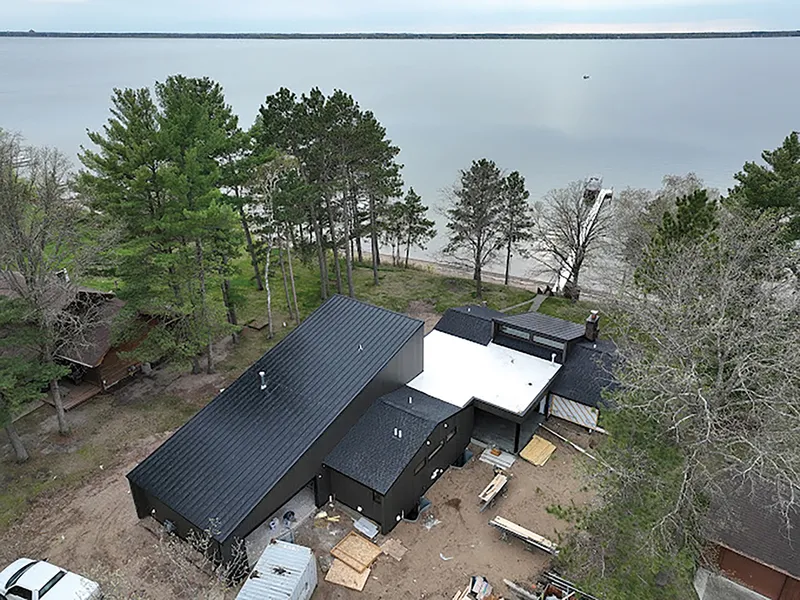 Aerial view of a dark modern home under construction near a sandy lakeshore with tall pines