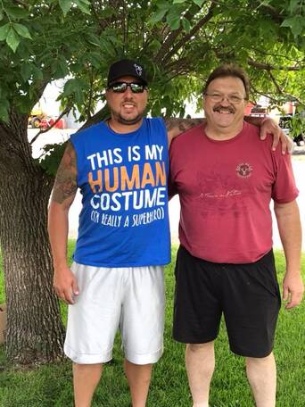 Two men smiling under a tree, one wearing a blue 'This Is My Human Costume' novelty shirt