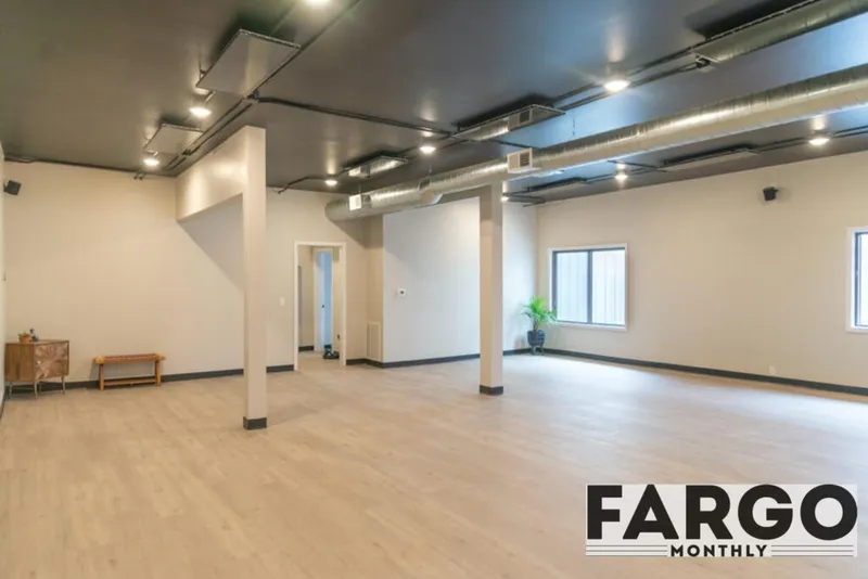 Spacious empty yoga studio with light wood floors, dark ceiling, exposed ductwork, and two windows