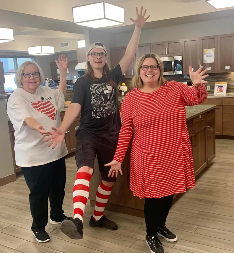 Two women and a young man pose with hands raised in a kitchen, wearing red and white outfits