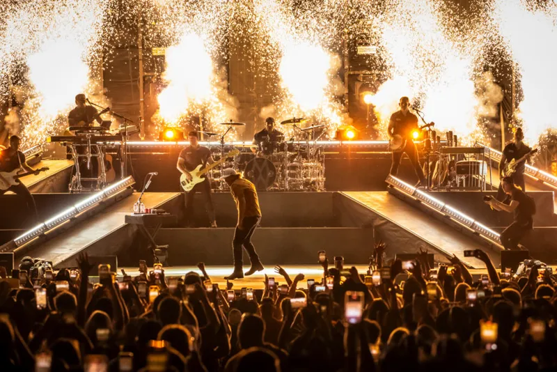 Band performs on stage with pyrotechnic fire bursts as crowd holds up phones