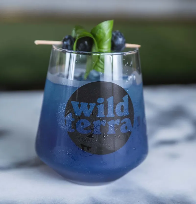 Blue cocktail garnished with blueberries and basil in a branded stemless glass