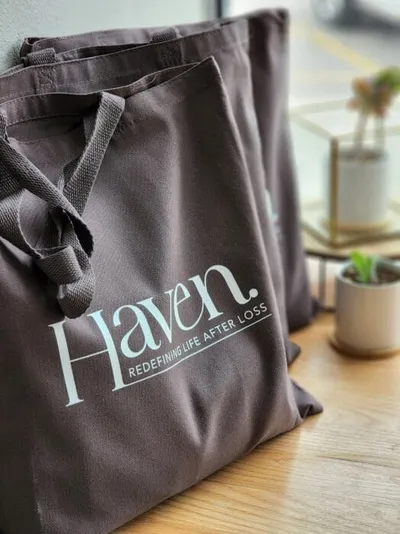 Dark gray tote bag printed with "Haven. Redefining Life After Loss" on a wooden surface