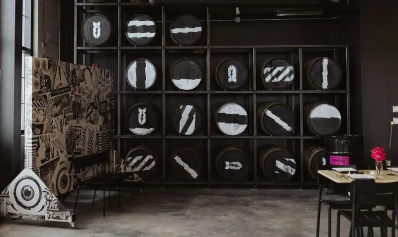 Dark industrial space with whiskey barrels displayed on metal shelving beside a large black-and-white illustrated panel