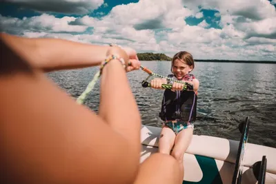 Young girl in a life jacket gripping a rope on a boat, smiling on a sunny lake
