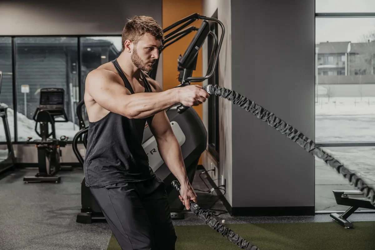 Muscular man in a black tank top doing battle rope exercises in a modern gym