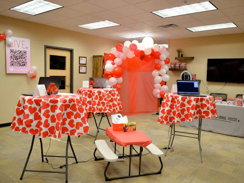 Indoor event room with red-and-white heart-print tablecloths, laptops, and a red-and-white balloon arch backdrop