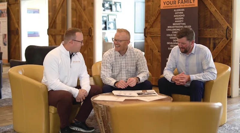 Three men laughing together in yellow armchairs inside a rustic office lobby