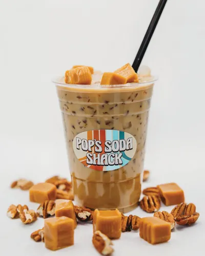 Pop's Soda Shack iced drink topped with caramel cubes and a black straw, surrounded by caramel candies and pecans