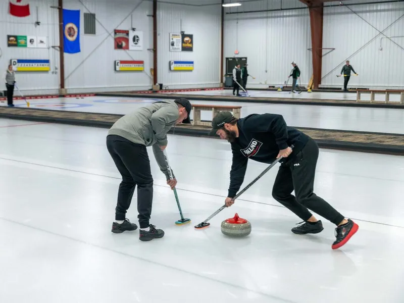 Two curlers sweep the ice beside a granite stone in an indoor curling rink