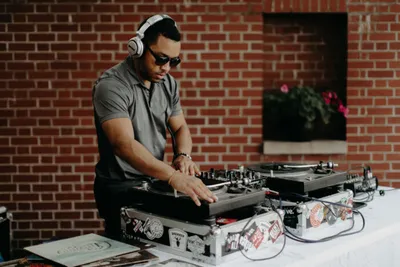 DJ wearing sunglasses and headphones working turntables outdoors against a brick wall