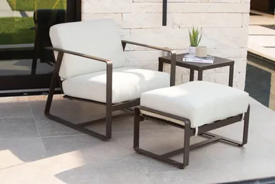 Dark bronze-framed patio lounge chair and ottoman with white cushions beside a small side table