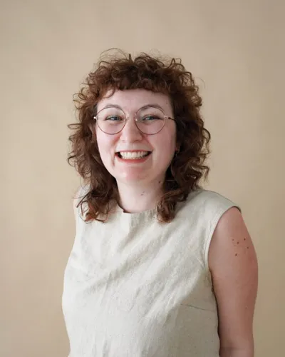Smiling woman with curly red hair and round glasses wearing a sleeveless sage green top
