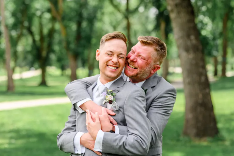 Two grooms in matching gray suits laughing and embracing outdoors among green trees