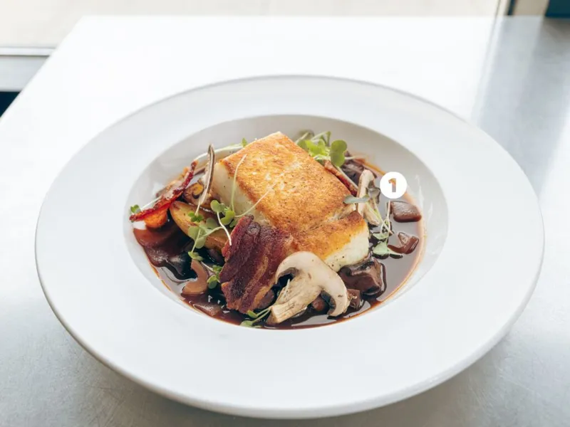 Seared white fish over mushrooms, bacon, and dark broth on a white plate