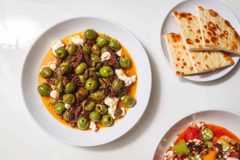Green olives with white cheese and red sauce on a plate, beside grilled flatbread and a tomato salad
