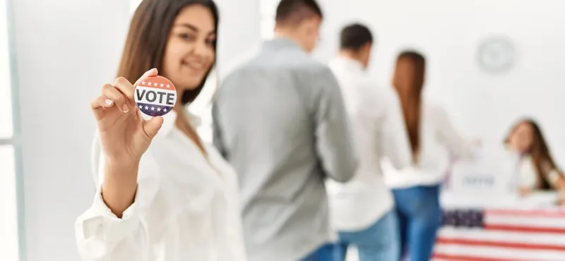 Smiling woman holding a red, white, and blue VOTE button inside a busy polling place