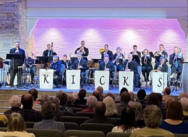 Big band ensemble labeled KICKS performing on a purple-lit stage before a seated audience