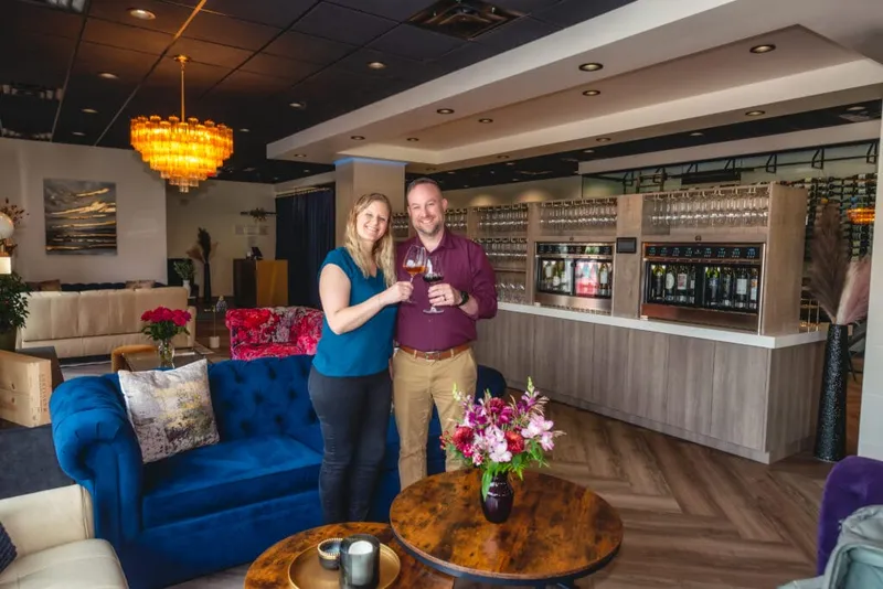 A smiling couple toasting wine glasses inside an upscale lounge with a tasting bar and blue velvet sofa
