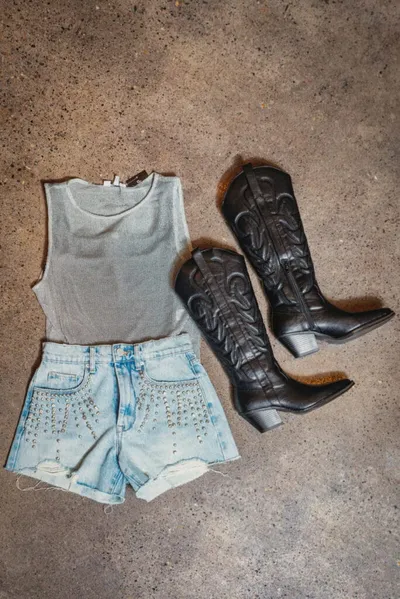 Flat lay of a gray crop top, studded denim cutoffs, and black cowboy boots on concrete
