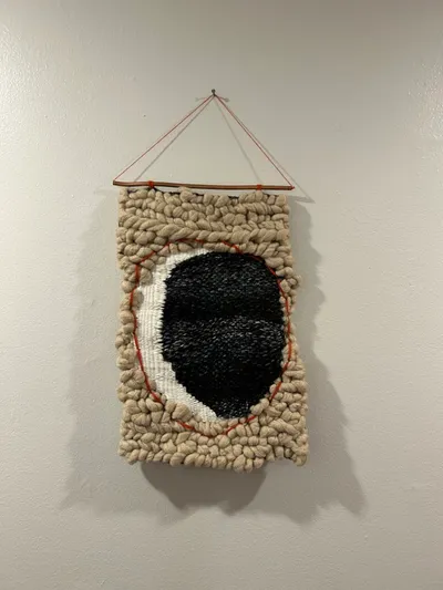 Chunky hand-woven wall hanging with cream yarn and a black-and-white oval center, trimmed in red, suspended from a wooden