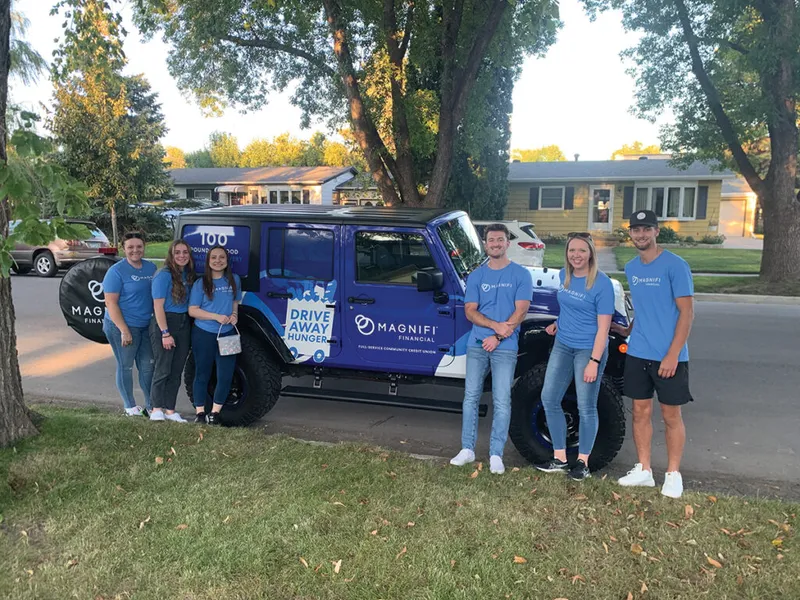 Five people in blue Magnifi Financial shirts posing beside a branded Jeep with Drive Away Hunger wrap