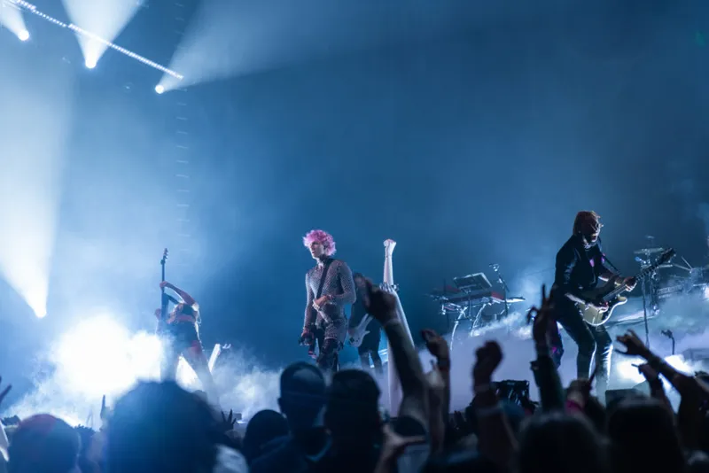 Pink-haired performer with guitarists on a fog-filled stage under blue spotlights, crowd arms raised