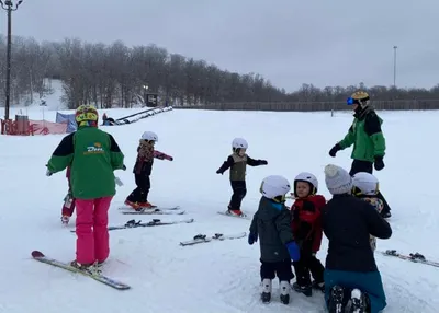 Two ski instructors in green jackets guide a group of young children on a snowy beginner ski slope