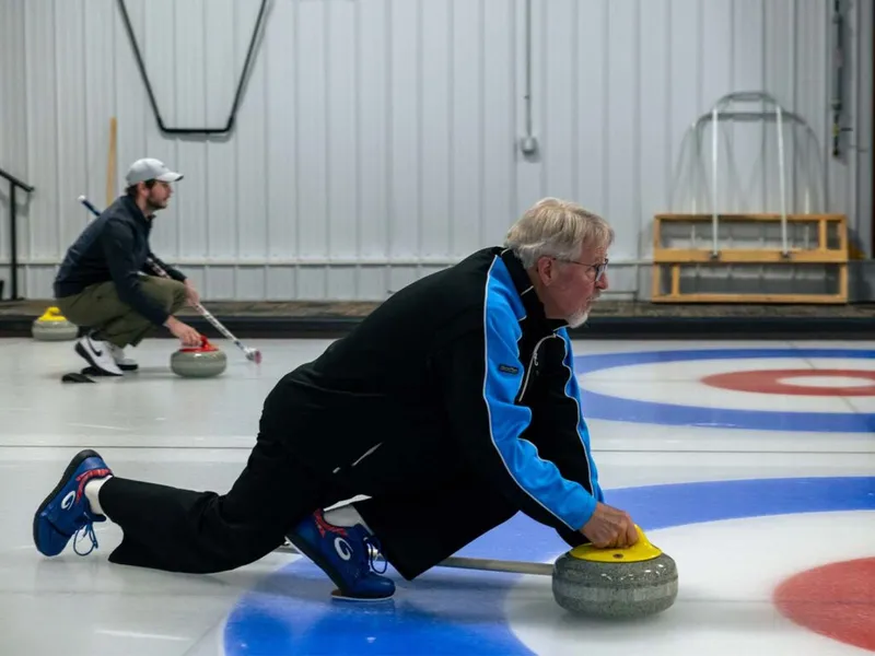 Older man in a blue jacket releases a curling stone on ice while another player crouches in the background