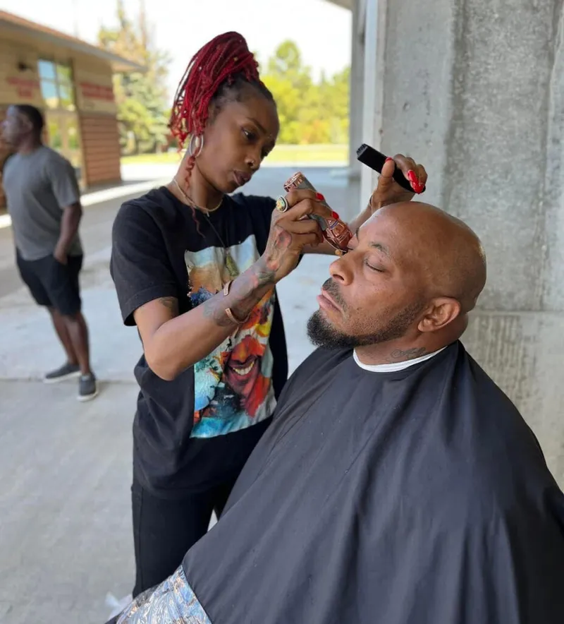 Woman with red braids uses clippers to trim a seated man's eyebrows outdoors