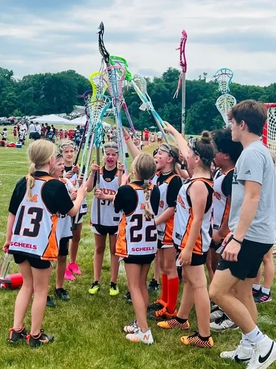 Youth girls lacrosse team in black-and-orange jerseys raising sticks together in a team huddle